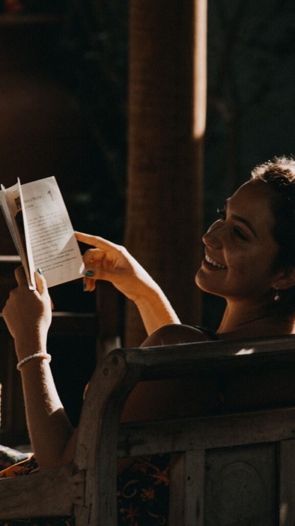 Smiling woman enjoys reading a book outdoors under warm sunlight.