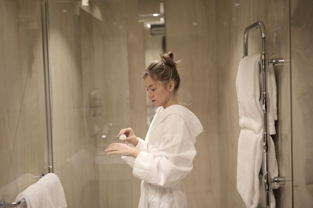 A woman in a bathrobe applies lotion in a contemporary bathroom with glass doors and towels.