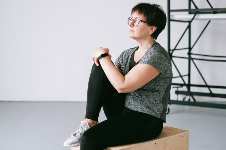Senior woman in active wear sitting on a wooden box indoors, reflecting on wellness.