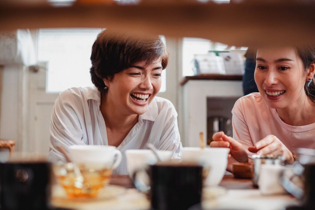 Cheerful young Asian female costumers in casual clothes laughing while choosing ceramic cups in modern shop