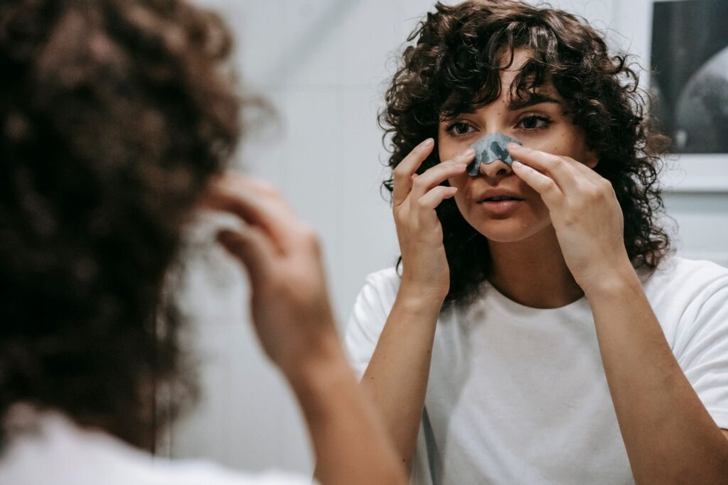Focused female with curly hair looking at mirror and applying clay mask on nose while standing in bathroom during daily routine