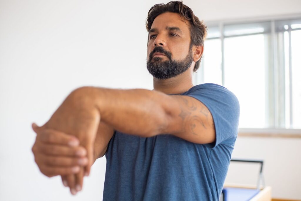 Man with beard stretching his arms indoors, practicing fitness routine.