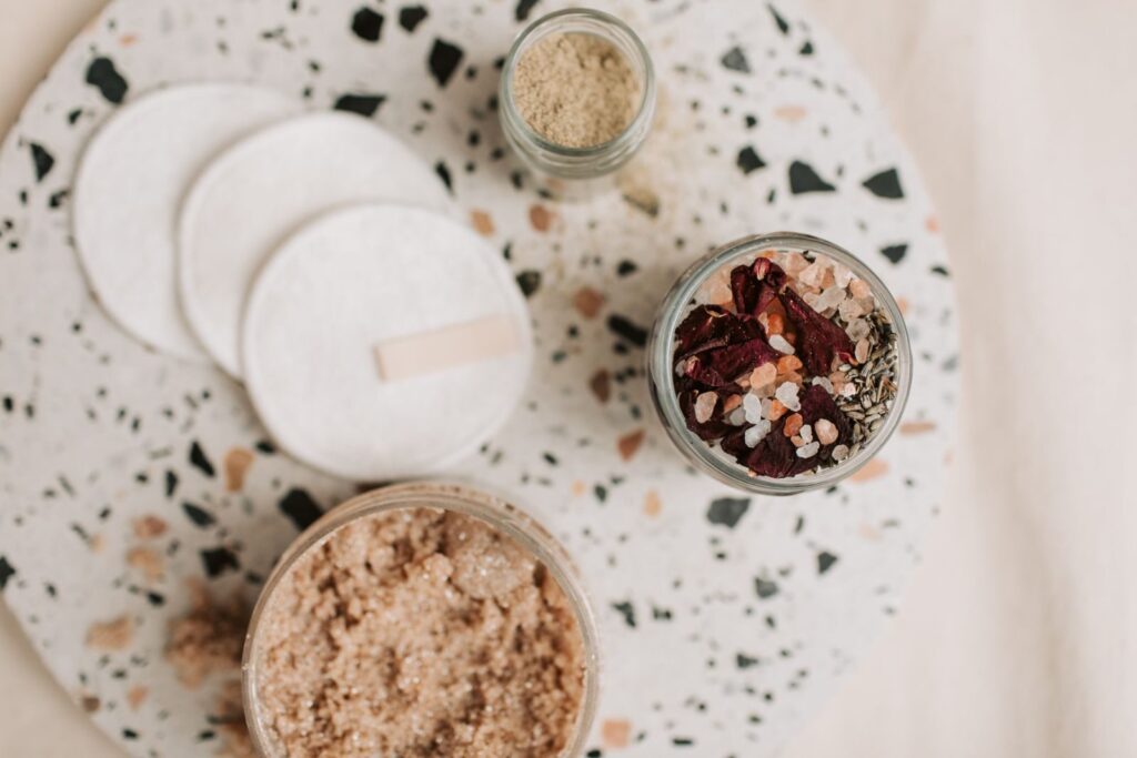 Flat lay of natural bath essentials including scrub and cotton pads on a terrazzo surface.