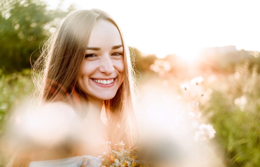 A young woman smiling brightly in a sunlit field during the golden hour.