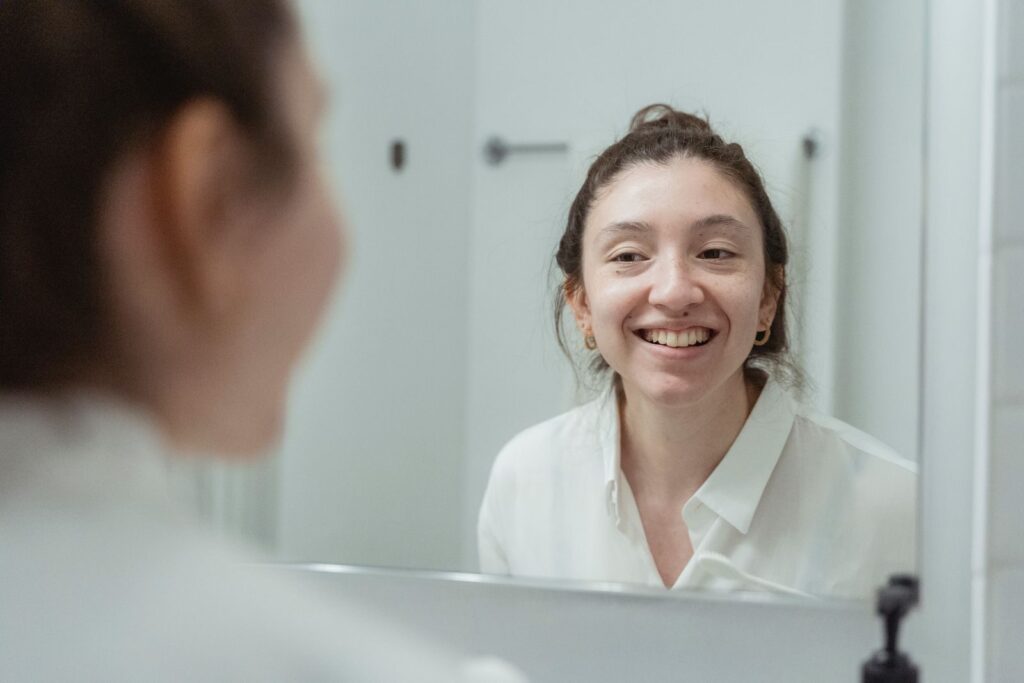 A woman in a white shirt smiles warmly at her reflection in a bathroom mirror.