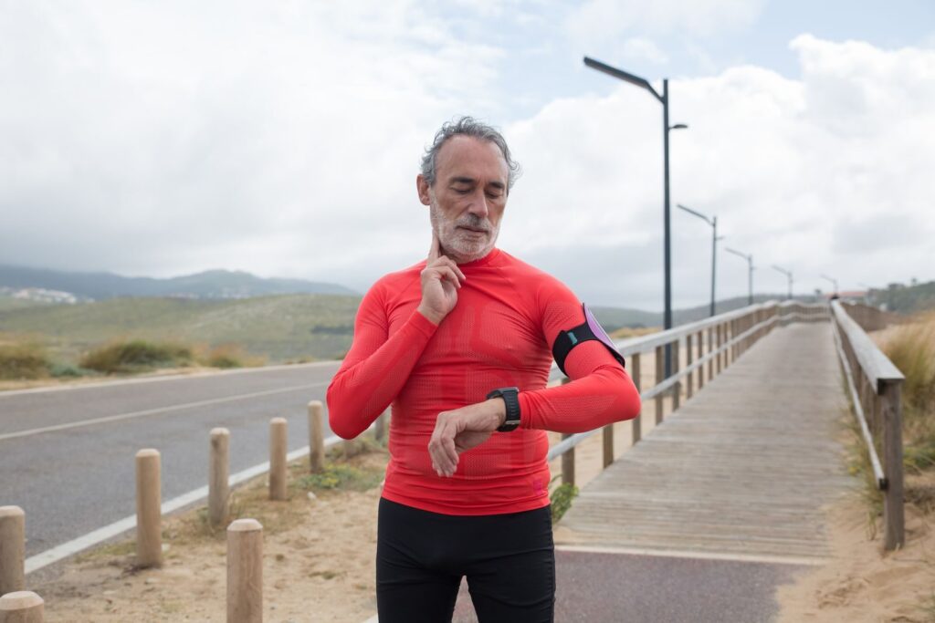 Elderly man in red activewear checking pulse on a coastal jogging path.