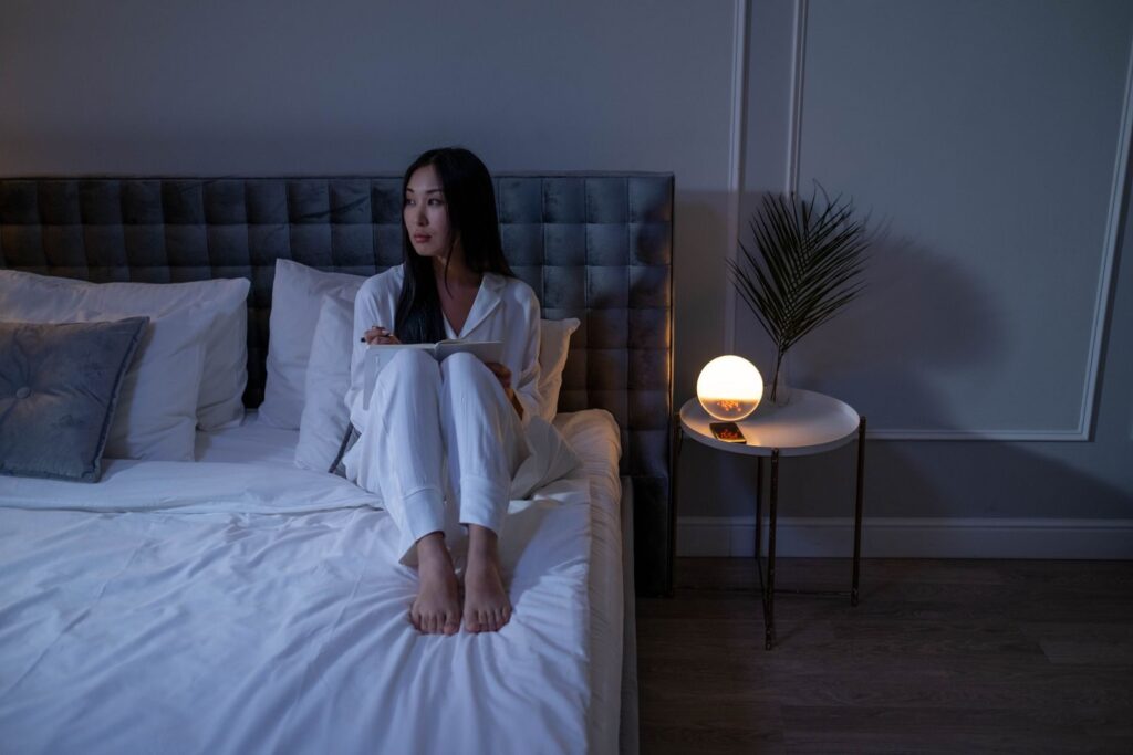 7 Astuces Pour Se Réveiller Plus Facilement le Matin | Santé et bien-être A woman in pajamas sits on a bed in a dimly lit bedroom, creating a serene nighttime atmosphere.