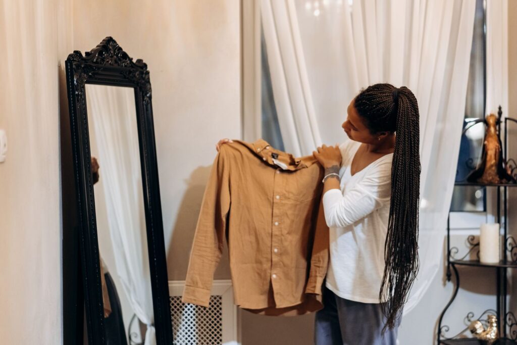 7 Astuces Pour Se Réveiller Plus Facilement le Matin | Santé et bien-être A woman examines a brown shirt in front of a mirror while shopping online at home.