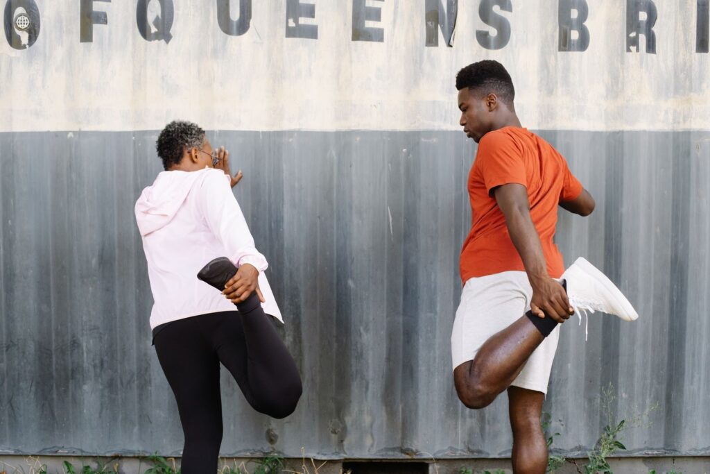 7 Étirements Pour Améliorer la Mobilité du Genou | Santé et bien-être Two adults stretching outdoors with 'Queens Bridge' sign in background.